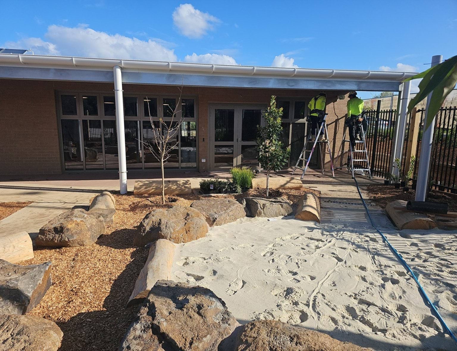 Newly landscaped courtyard of Biyala Primary School with tanbark, large stone and logs surround a sandpit. Two tradespeople are on ladders in the background. 