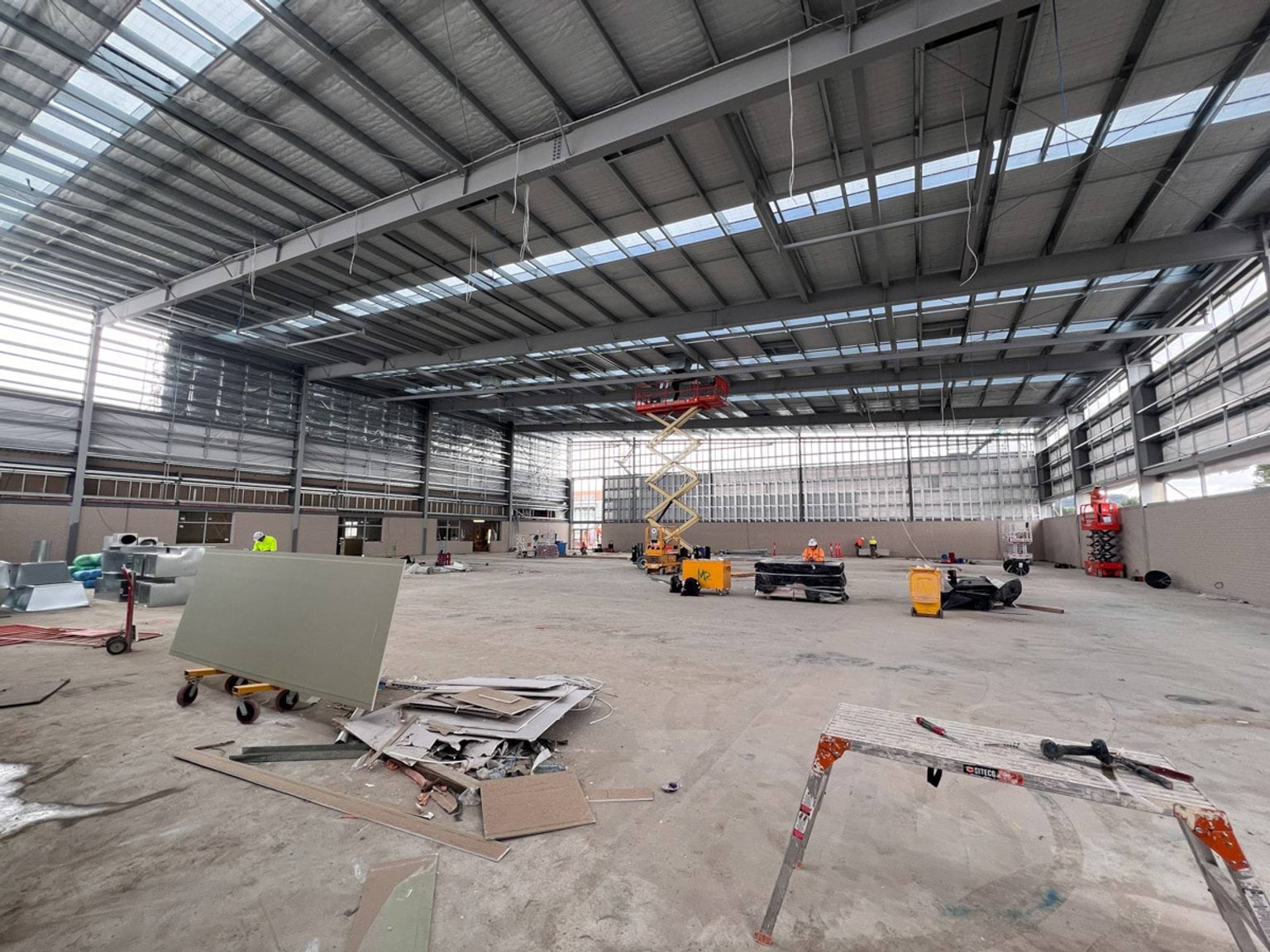 Scissor lift extended to the ceiling of an auditorium under construction. Leftover large cladding is stacked in a nearby trolley. 