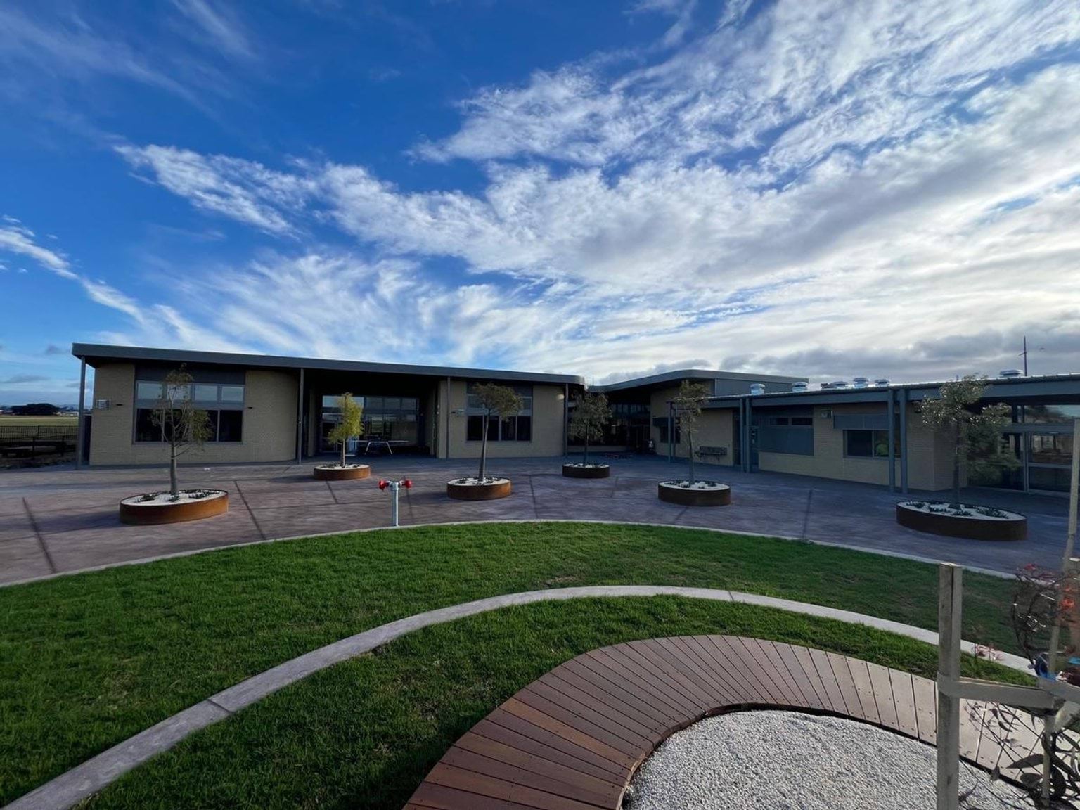 Raised garden beds have been placed in a zig zag pattern in a concreted area of the playground. Rounded landscaping features with grass wrapping around timber path
