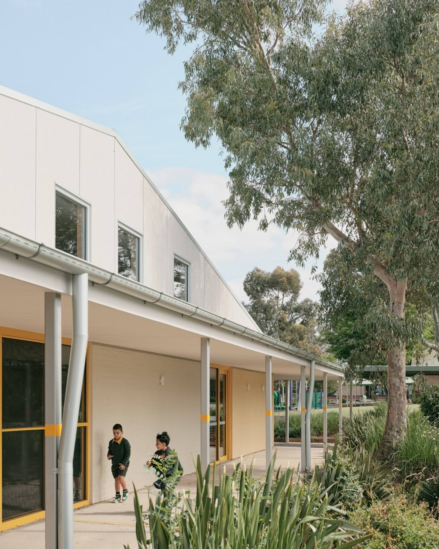 The exterior of Banyan Fields Primary School, showing the verandah along the side of the building. Two kids are running past. There is a tall tree on the right side. 