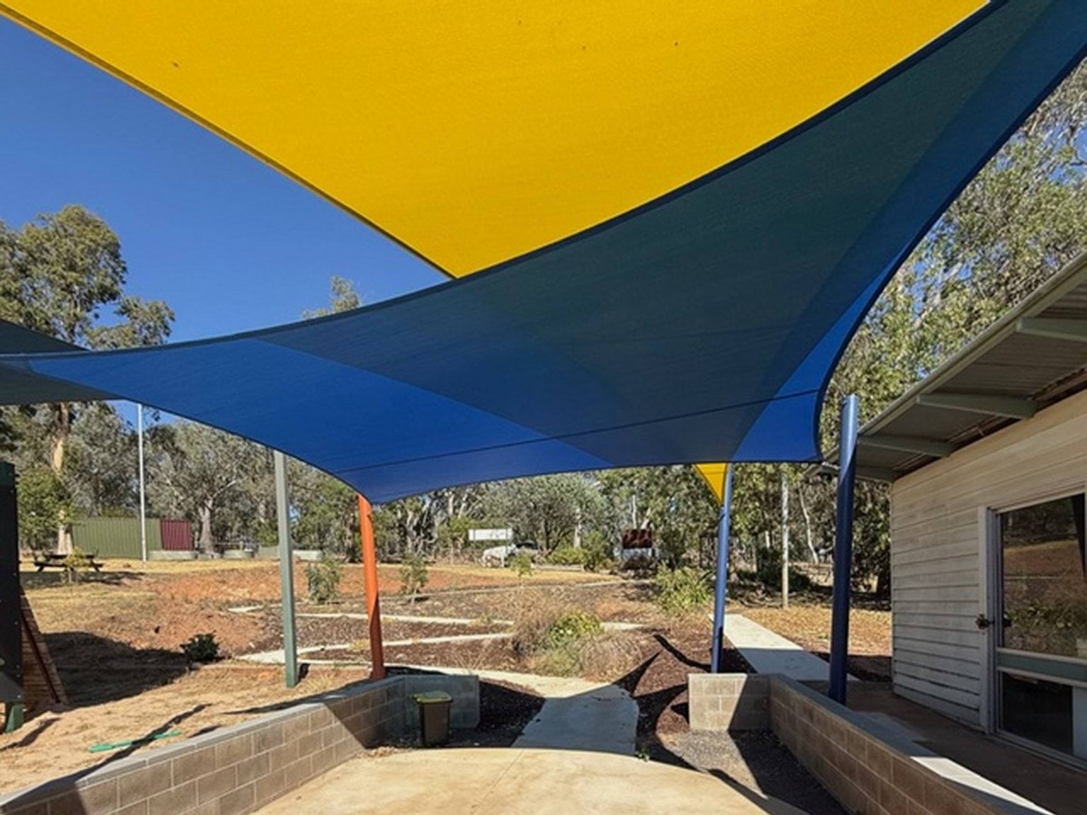 Outdoor photo of Currawa Primary School. It shows a shaded seating area, with blue and yellow shade cloths. There is a path leading to the background.