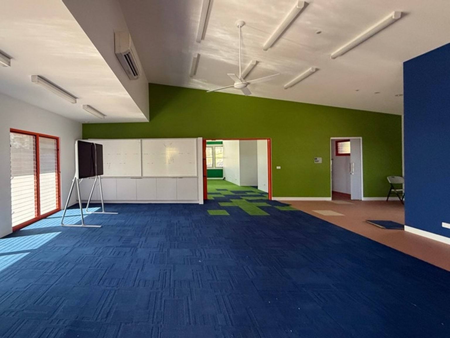 Indoor photo of a school building. There is a screen at the front of the room, carpeted floors, a ceiling fan, and blue and green walls.