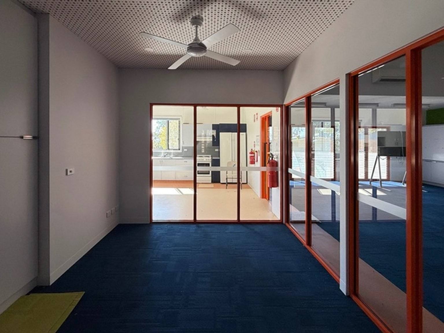 Indoor photo of a room with doors on either side. There is a blue and green carpet, ceiling fan, and kitchen area in the background.