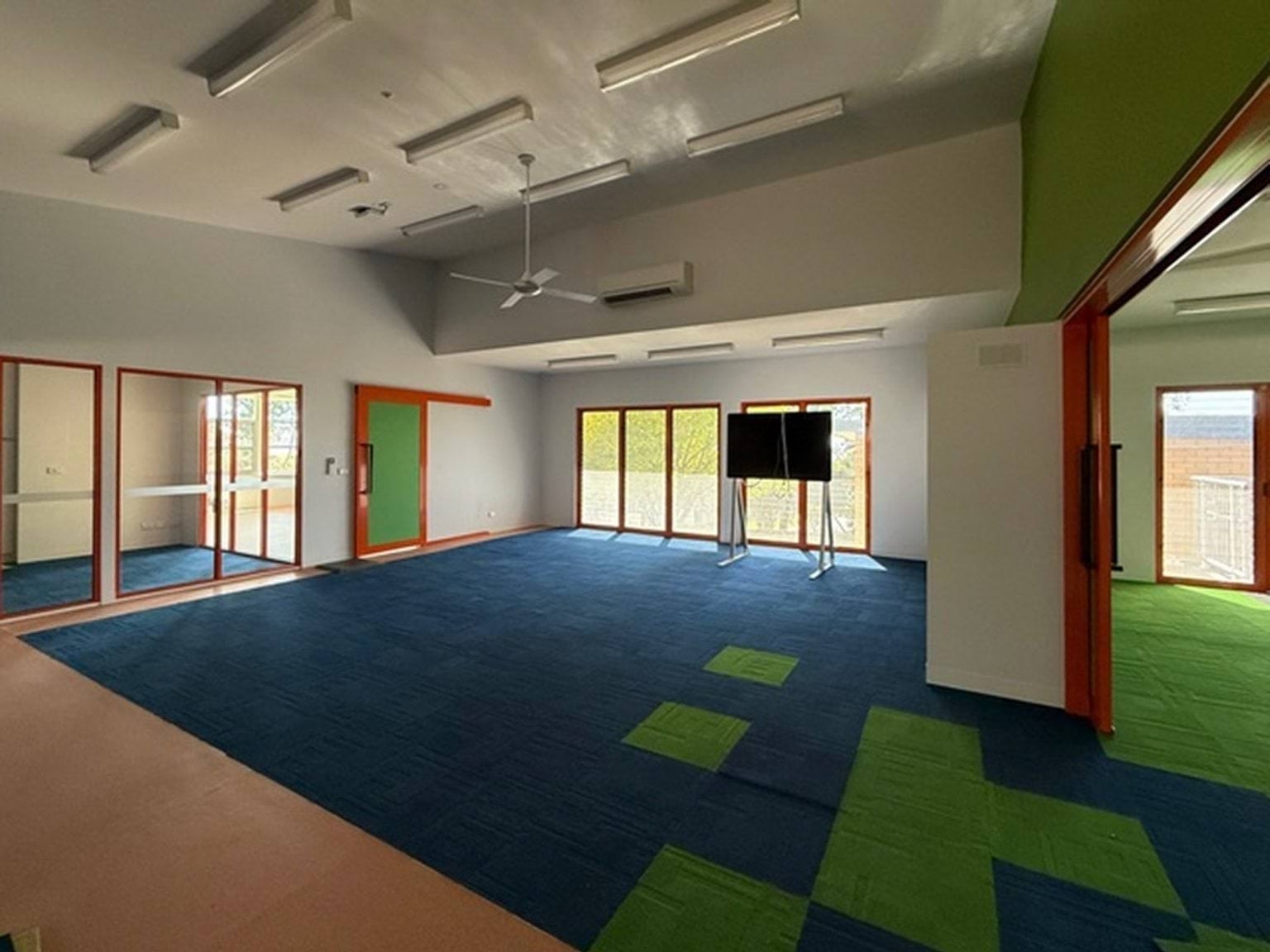 Inside photo of a school room. It has an orange, blue and green carpet with a modern design. There are doors that open onto rooms on either side. There is a board at the front of the room.