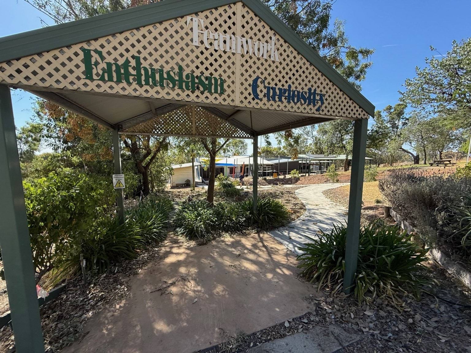 Outdoor photo of a wooden gazebo. It is green with cream coloured lattice, and has signage which reads "teamwork, enthusiasm, curiosity". 