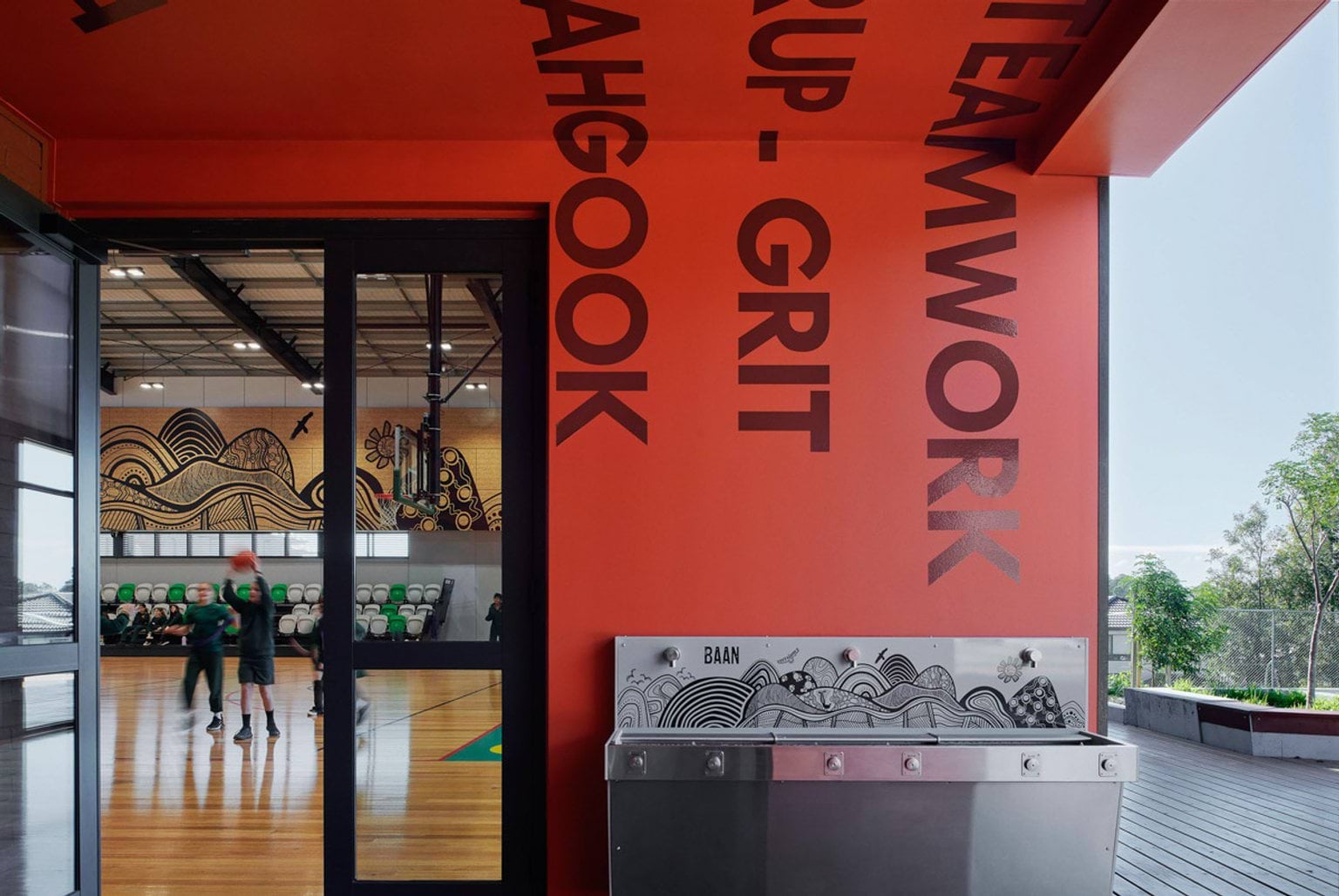 The entrance to the gymnasium at Greenhills Primary School. The wall and roof leading in are painted red, with words painted on in big letters. There are also drinking fountains with an artwork behind them. Looking into the gym there are students playing basketball on a hard court.