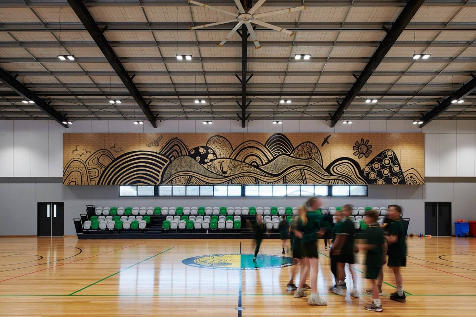 Inside the gymnasium at Greenhills Primary School. There are students in a huddle on the hard court. Behind them are the stands with green and white seats, and a big artwork hanging on the wall.