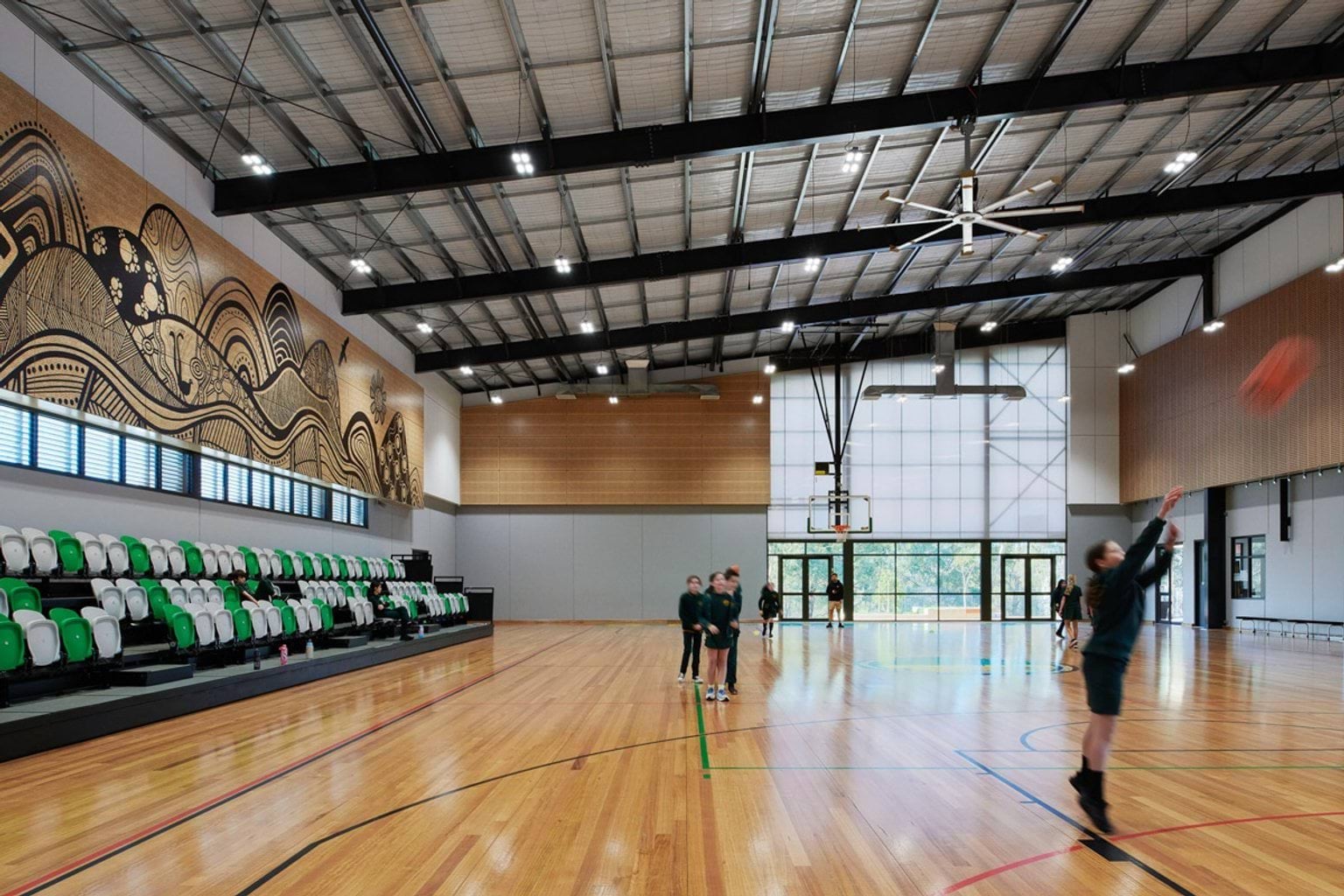 Inside the gymnasium at Greenhills Primary School. There are students moving around the hard court playing basketball. On the left are the stands with green and white seats, and a large artwork hanging on the wall above.