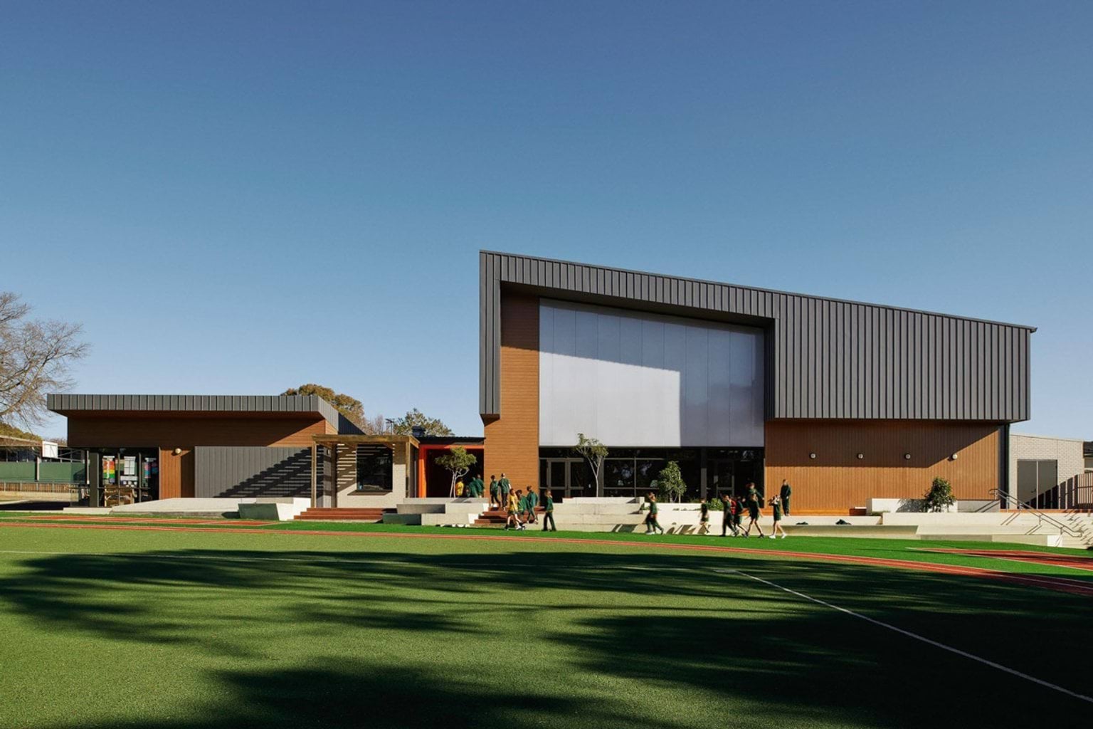 Outside Greenhills Primary School. There is a grassed field in the foreground, and buildings in the background with concrete tiered steps that students are walking past.