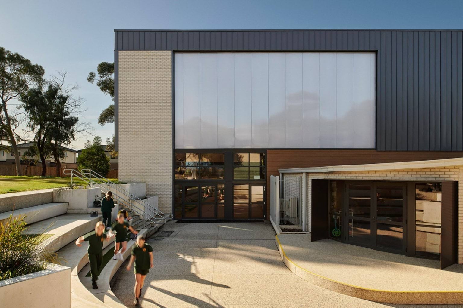 Outside Greenhills Primary School. Students are walking around the tiered concrete steps that lead down into the school and up to a grassed area.