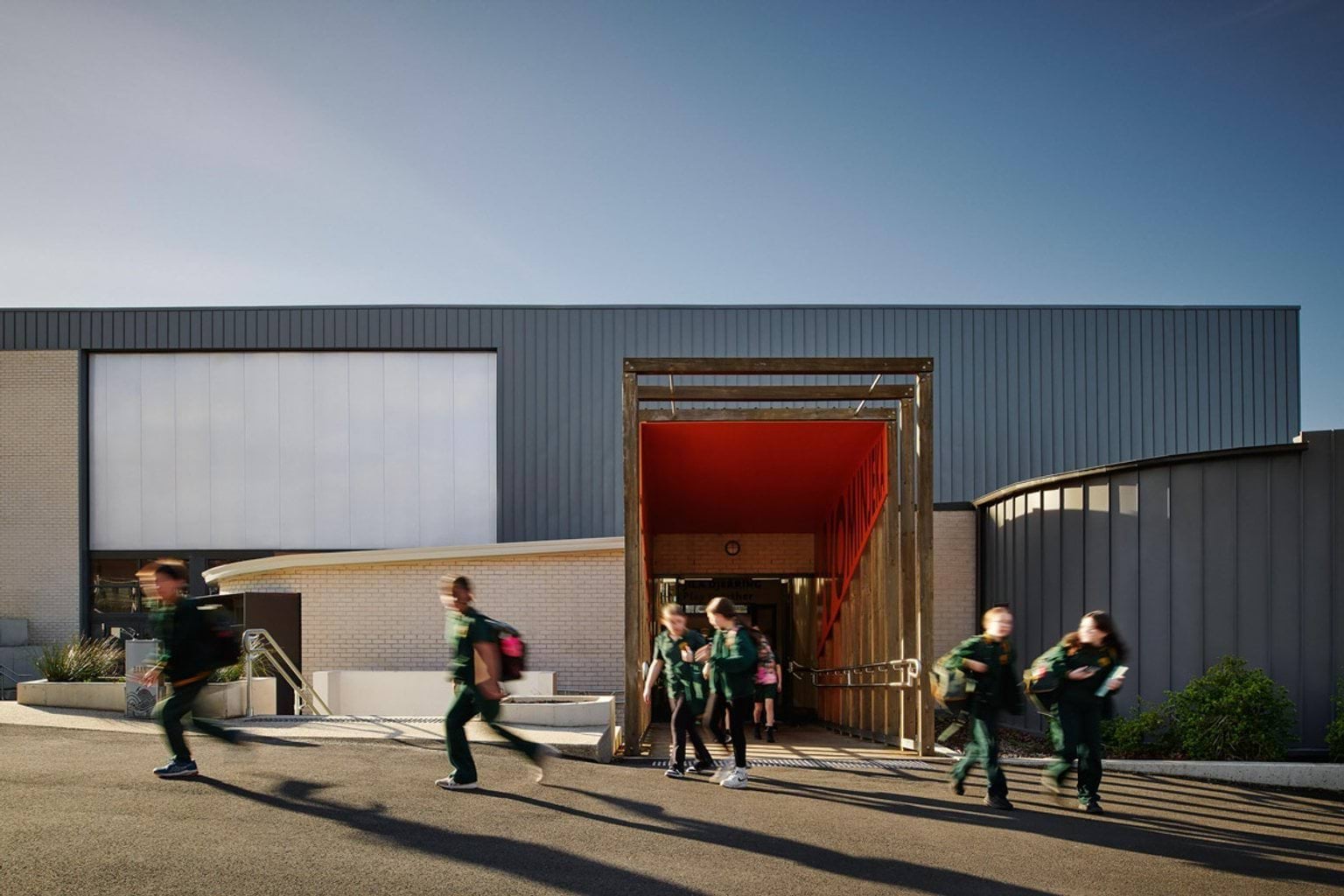 Showing an entry/exit into Greenhills Primary School, that is made from wooden square arches with a red banner wrapped on the inside. Students are walking out of it. The building is part cream brick and part blue-grey wood.