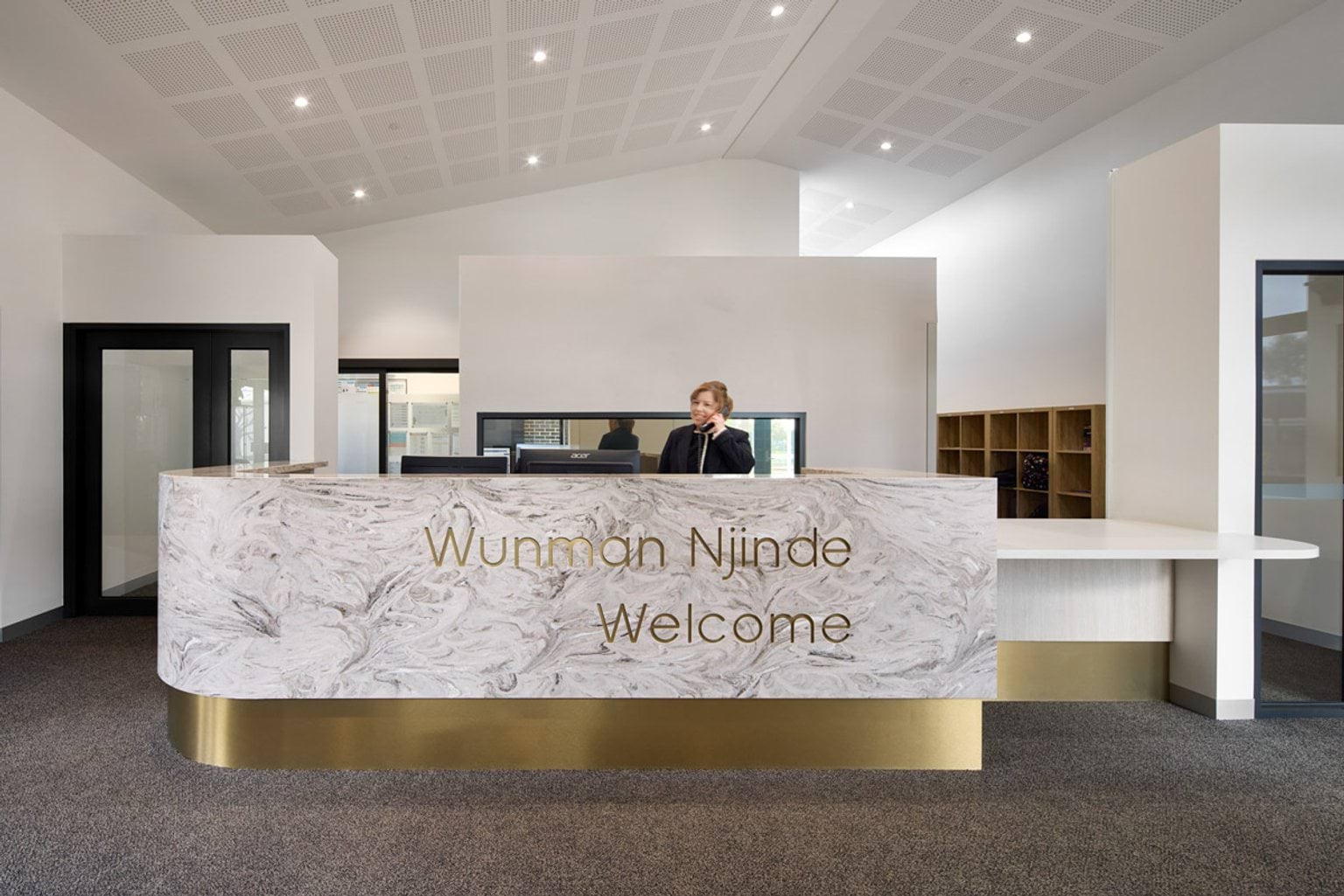 Indoor photo of Hazelwood North Primary School. It has a welcome desk with modern design and metallic gold trip. There is a person on the phone, and doors behind them leading to other parts of the building.