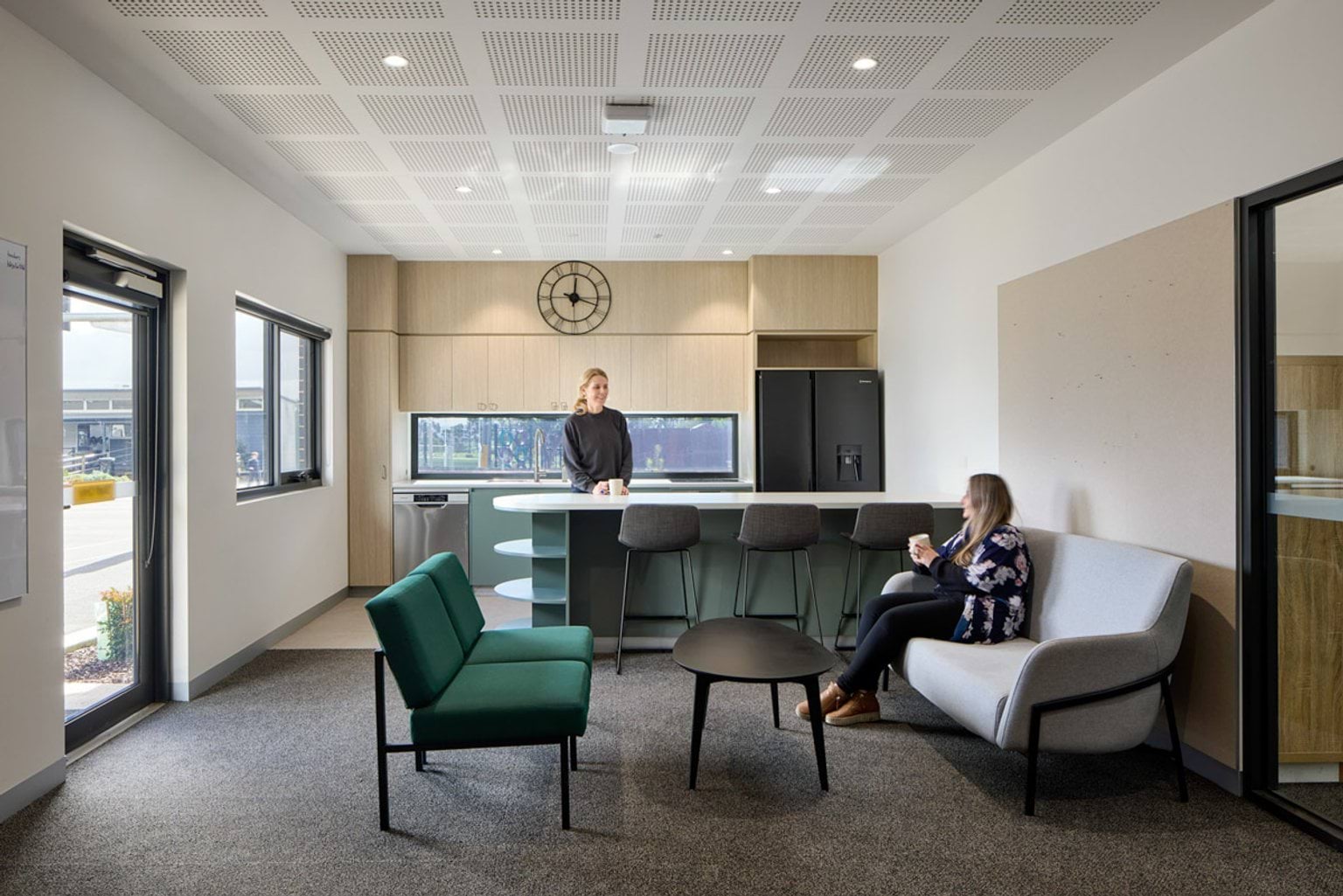 Indoor photo of Hazelwood North Primary School. It is a seated break area with kitchenette. There are two women holding coffee and talking. There is a bench with high seats, and a coffee table with lower seats. Windows face outside.
