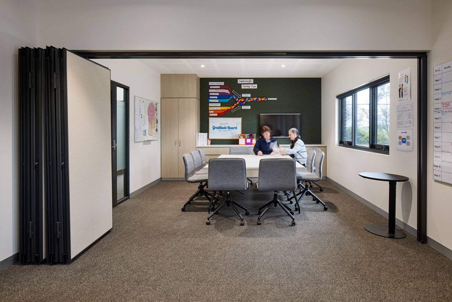 Indoor photo of Hazelwood North Primary School. There is a square table surrounded by chairs where two people are browsing notes. There is a green feature wall, cupboard, and whiteboard.