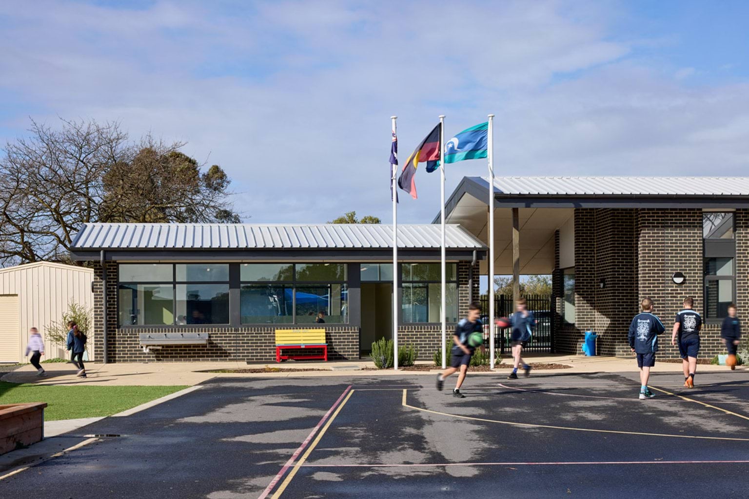 Outdoor photo of basketball court with school building in background. There are five children in school uniforms playing basketball.