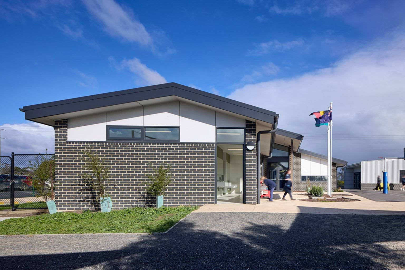 Outdoor photo of school building. It has grey brick walls, white panels, and a dark grey roof. There are paths leading to it and saplings next to the walls.