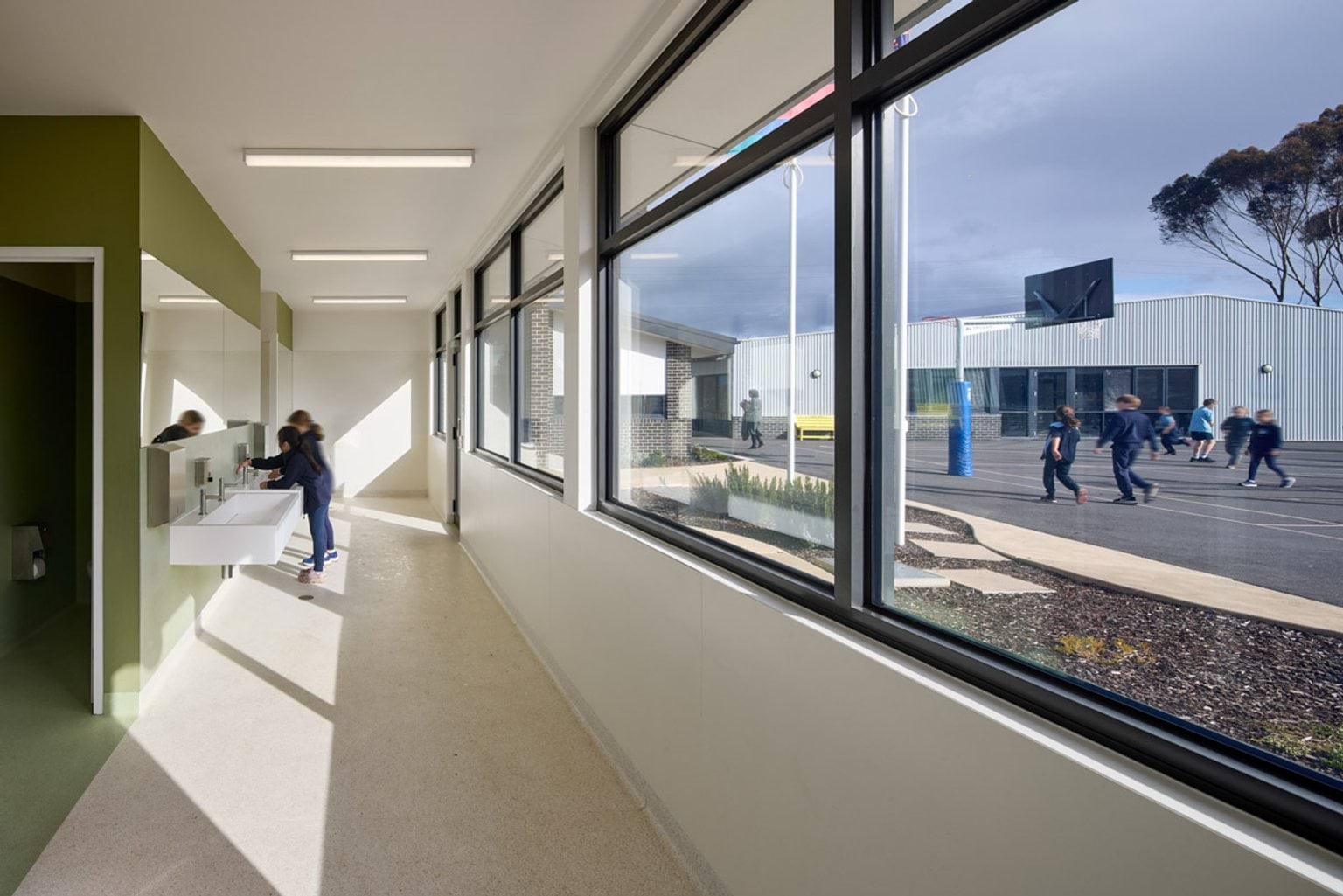 Indoor photo of school bathroom building. There are large windows on the righthand side through which a basketball court is visible. Inside, there are school students at a water tap.