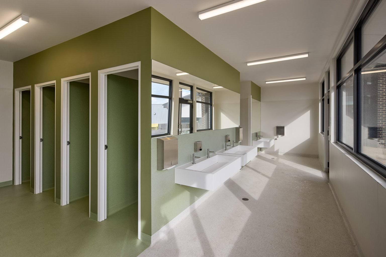 Indoor photo of school bathrooms. There are four stall doors on the left side, with basins around the corner on the right side. The walls are green with white accents.