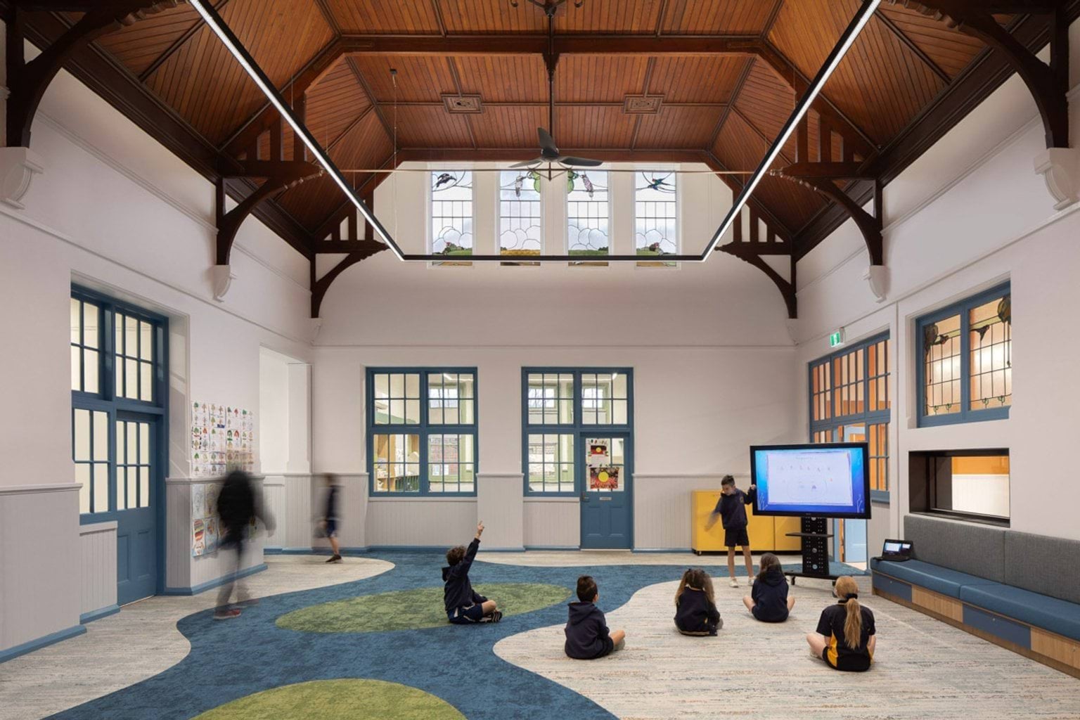 Inside a room at Moonee Ponds West Primary School. There is a very high wooden ceiling and white walls. There is a carpet with a colourful design. Kids are sitting on the carpet facing a big TV. There are lots of windows letting in light.