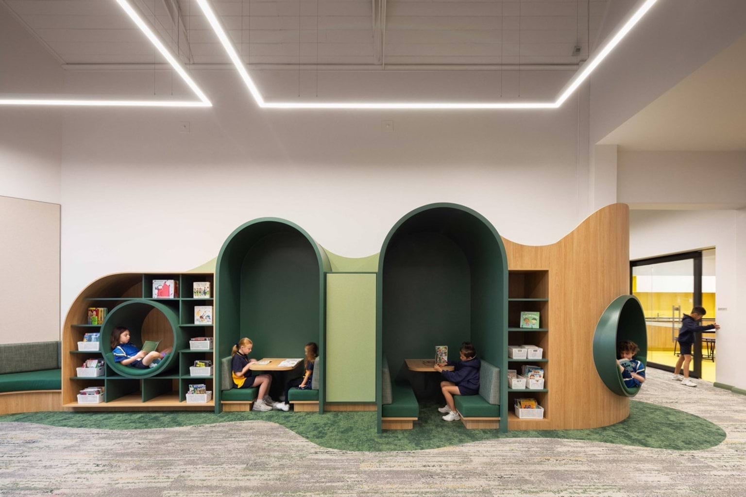 A wall at Moonee Ponds West Primary School that has recessed sections to sit in. 2 are circles where kids are sitting and reading, and 2 sections are arches with tables and bench seats. There are books and containers of equipment on shelving around these sections.