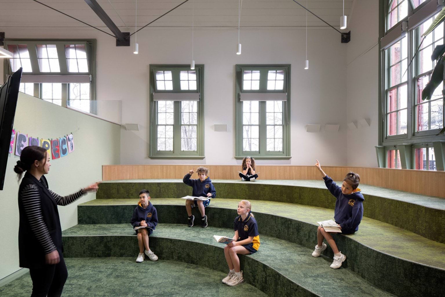 Inside Moonee Ponds West Primary School. The floor has green carpeted tiered steps that kids are sitting on, facing a teacher at the front. There are big windows with green window frames on the walls.