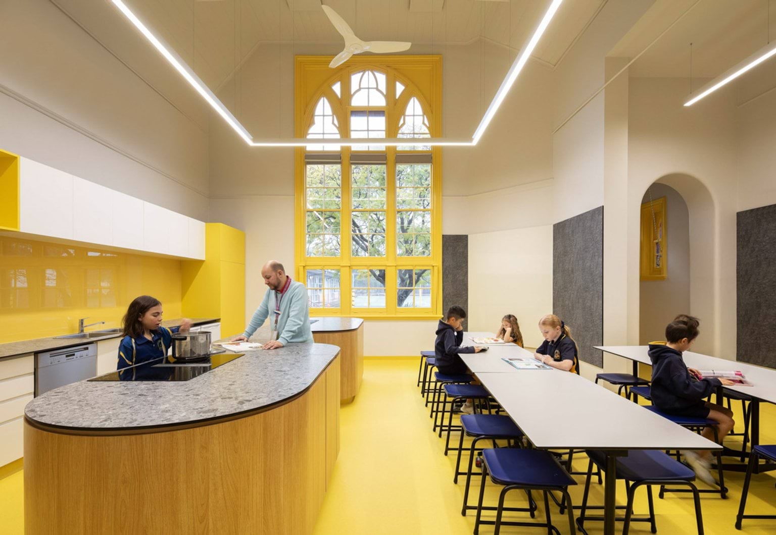 Inside an eating area at Moonee Ponds West Primary School. There are kids sitting at long tables on the right, and a kid is using a pot on the stove on the left. An adult stands near them. On the left-hand side wall is a sink and dishwasher. The room's floor, splashback and window frame are painted yellow.