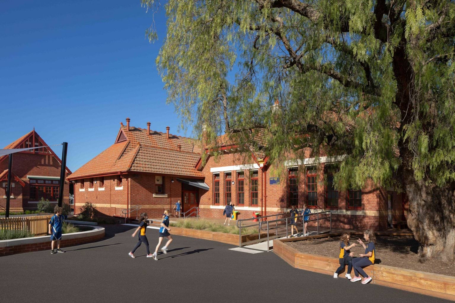Outside Moonee Ponds West Primary School. Kids are running around or sitting together and playing. The school building has a red brick exterior. There is a big tree with leaves in the right of the photo.