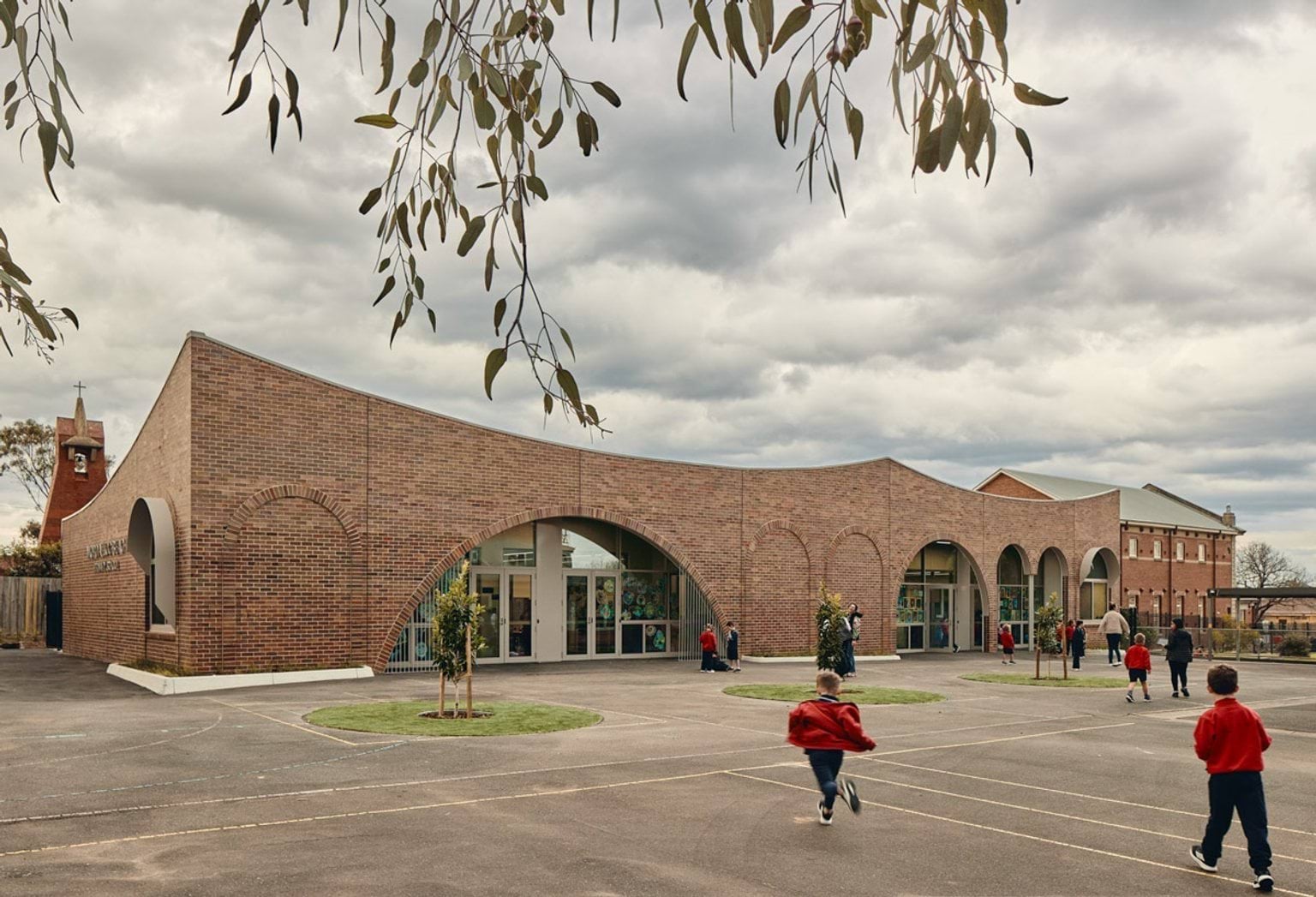 Outdoor photo of brick administration building. There are arches above the doors and windows, a concrete area outside, and saplings planted in grass circles. Children in school uniforms are walking in front of the building.
