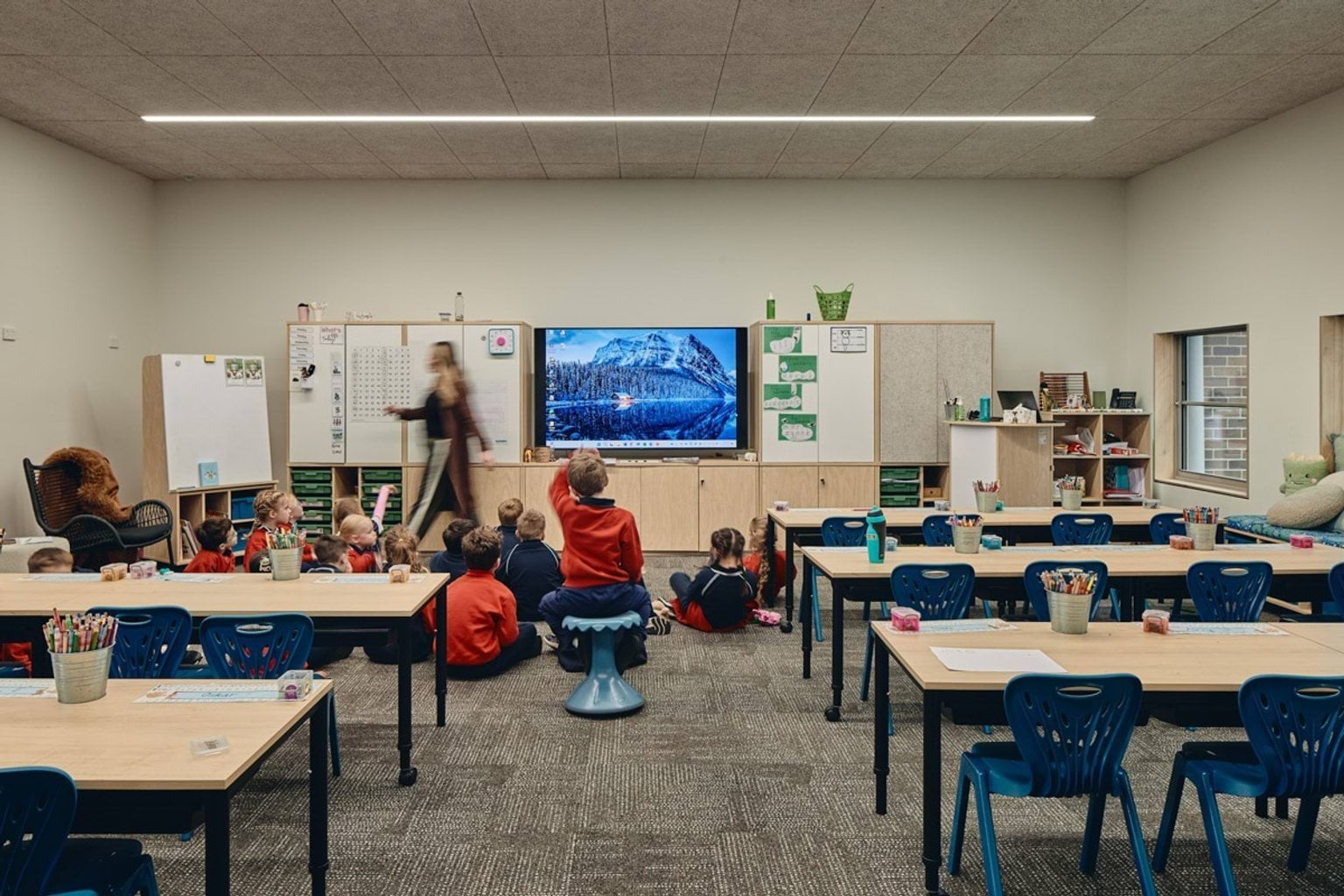 Indoor photo of classroom. There are five tables with blue seats behind them. At the front of the class, there is a screen, whiteboards, and a teacher standing in front of school students.