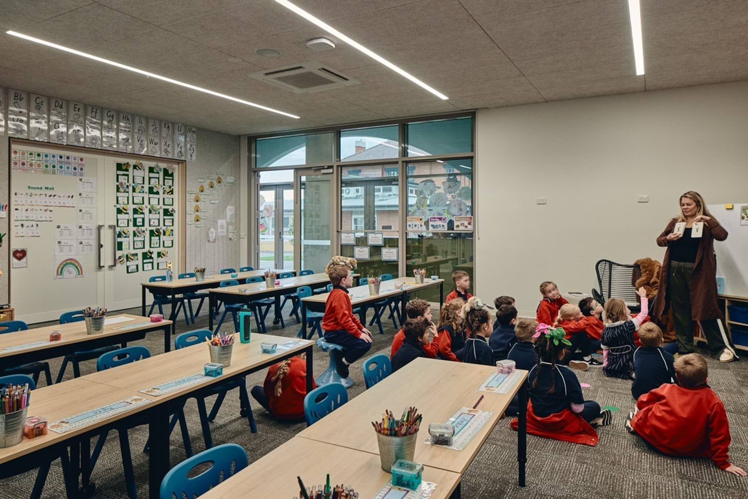 Indoors photo of classroom. There is a group of students sitting on a grey carpeted floor looking at a teacher who is standing in front of them. There are white boards, tables with chairs, and pictures on the walls.