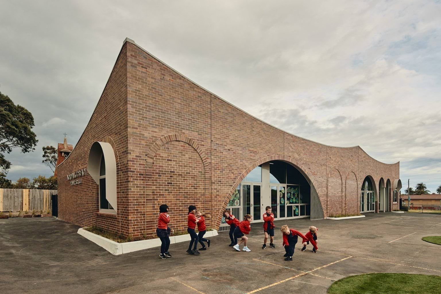 Outdoor photo of school administration building. It is a brown brick building with arches above its doors and windows. There is a group of children in school uniforms standing outside.