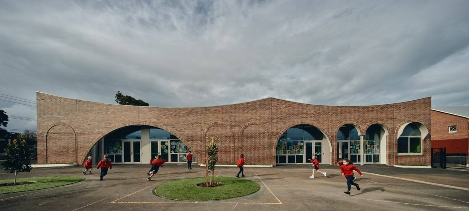 Outdoor photo of brick administration building. There are arches above the doors and windows, a concrete area outside, and saplings planted in grass circles. Children in school uniforms are walking in front of the building.