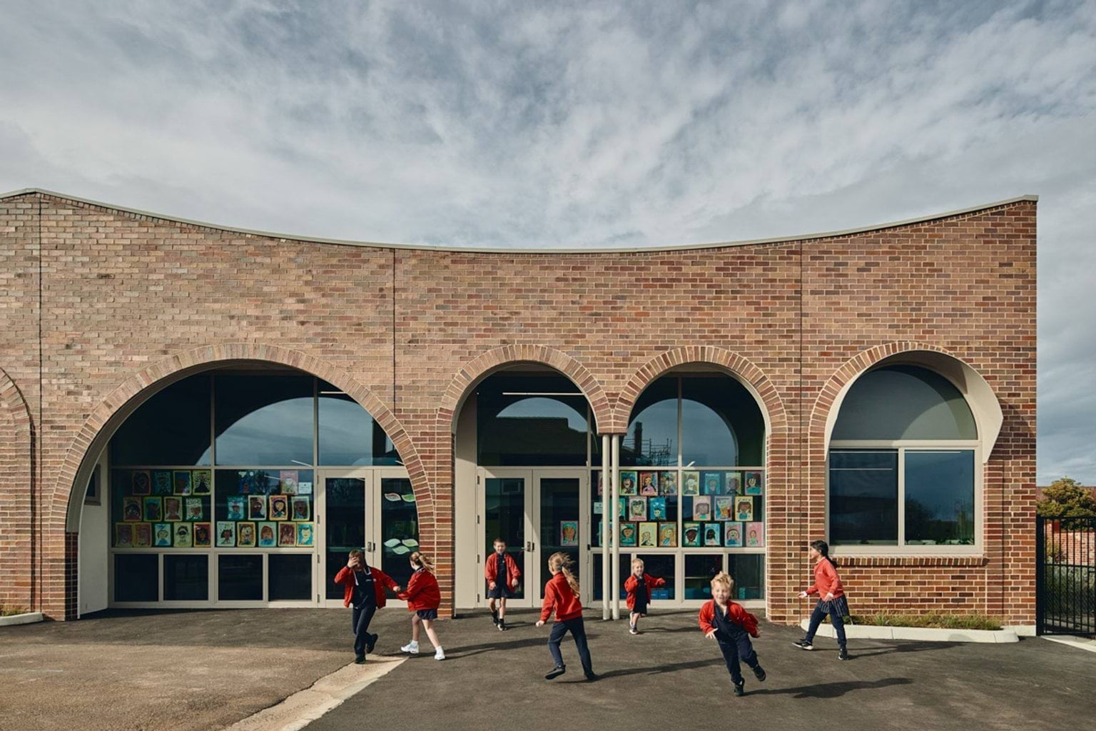 Outdoor photo of brick administration building. There are arches above the doors and windows, a concrete area outside, and saplings planted in grass circles. Children in school uniforms are walking in front of the building.