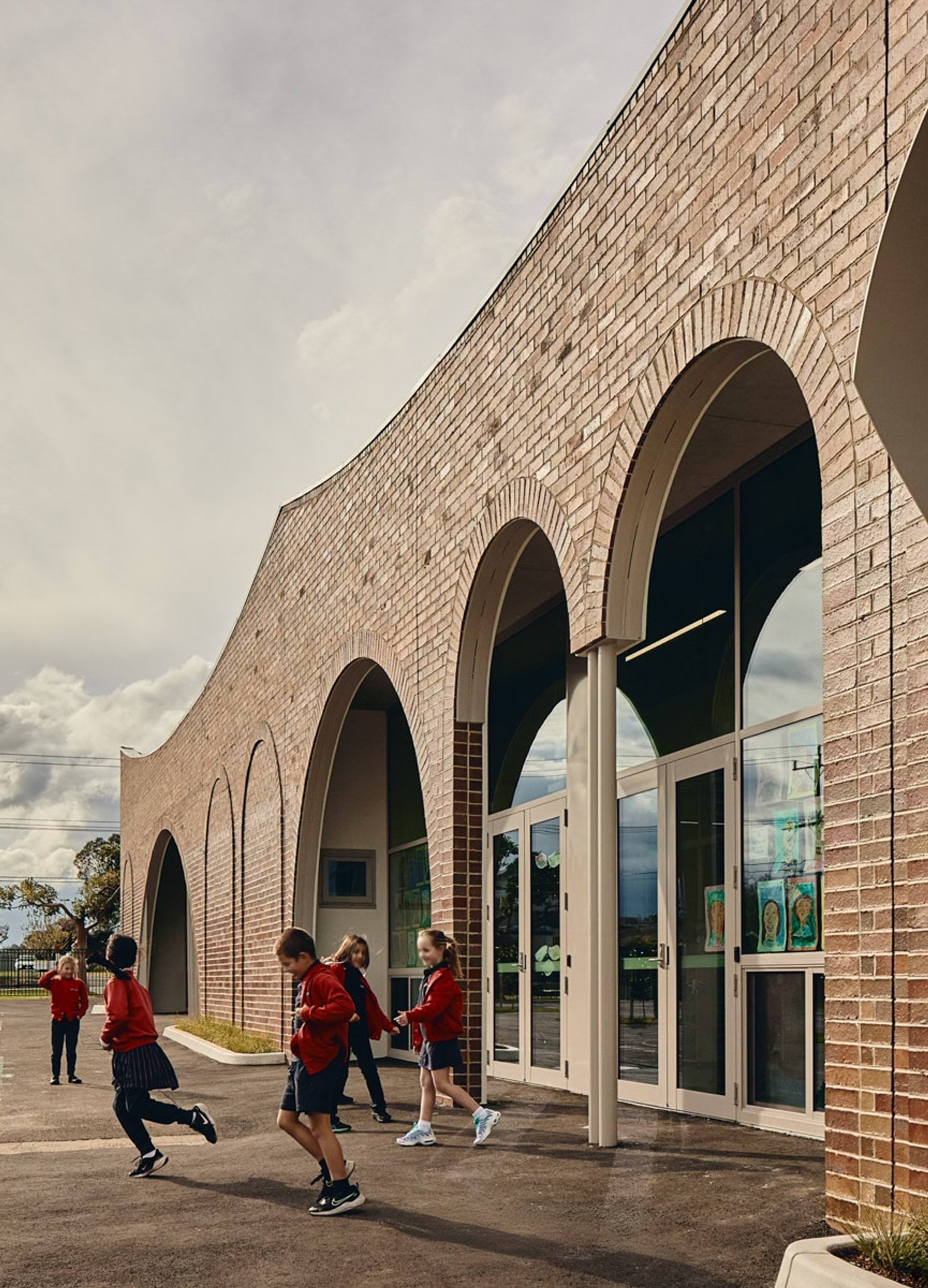 Outdoor photo of brick administration building. There are arches above the doors and windows, a concrete area outside, and saplings planted in grass circles. Children in school uniforms are walking in front of the building.