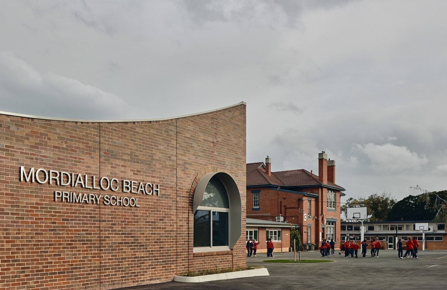 Outdoor photo of brick administration building. There are arches above the doors and windows, a concrete area outside, and saplings planted in grass circles. Children in school uniforms are walking in front of the building.