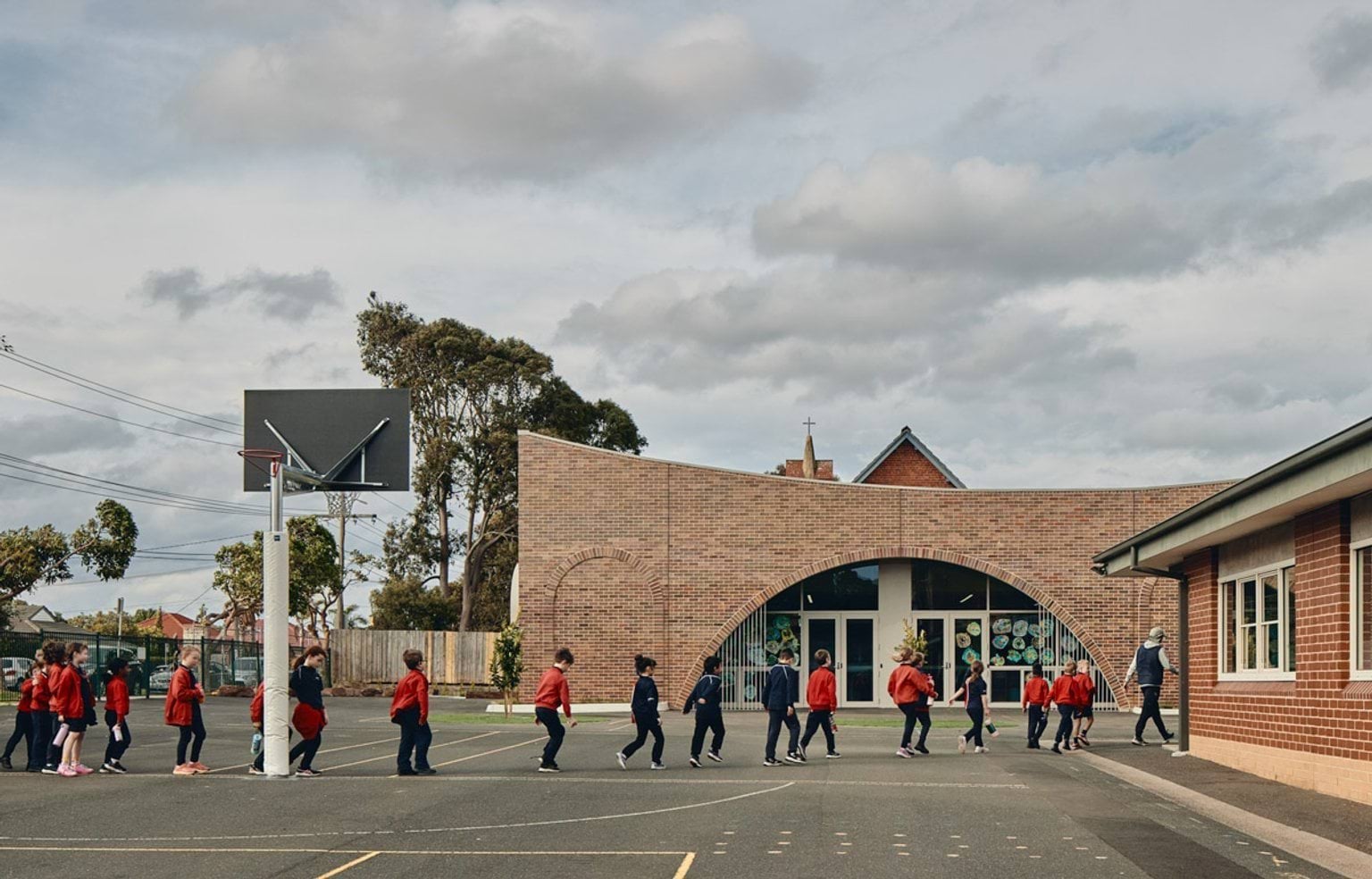 Outdoor photo of brick administration building. There are arches above the doors and windows, a concrete area outside, and saplings planted in grass circles. Children in school uniforms are walking in front of the building.
