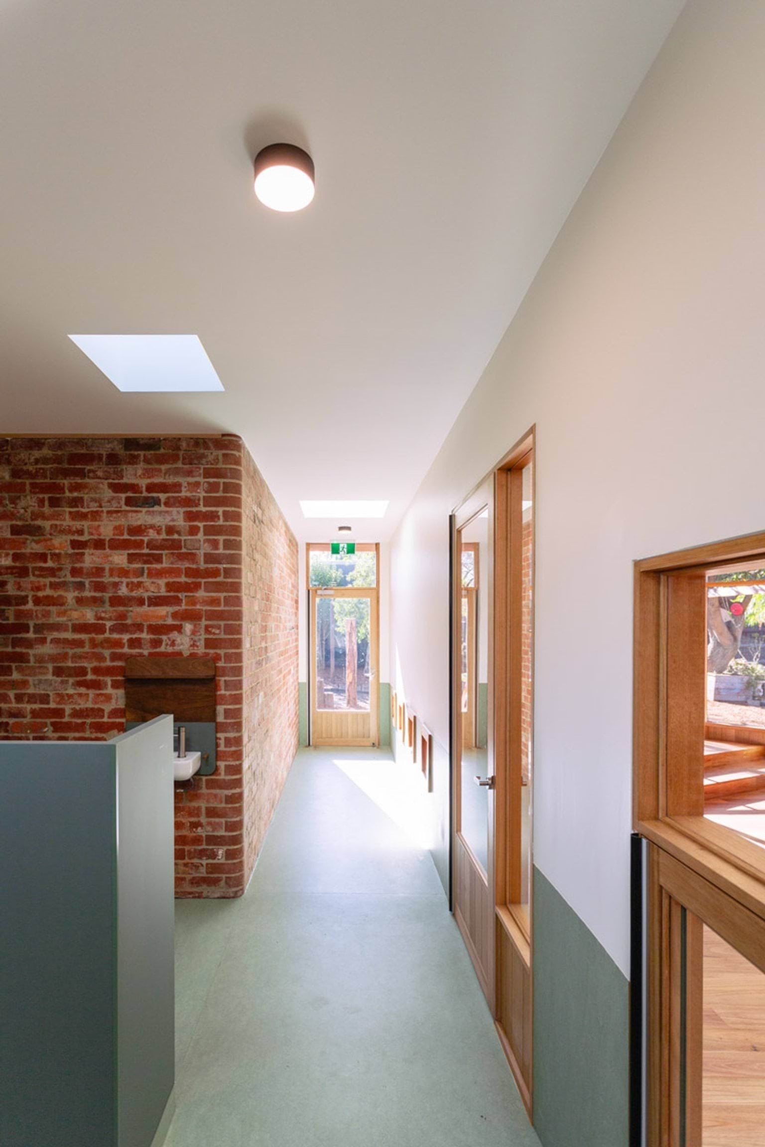 Inside a hallway of St Kilda & Balaclava Kindergarten. The ground is green with white walls and a brick wall. There is a door at the end of the hallway and on one side. 
