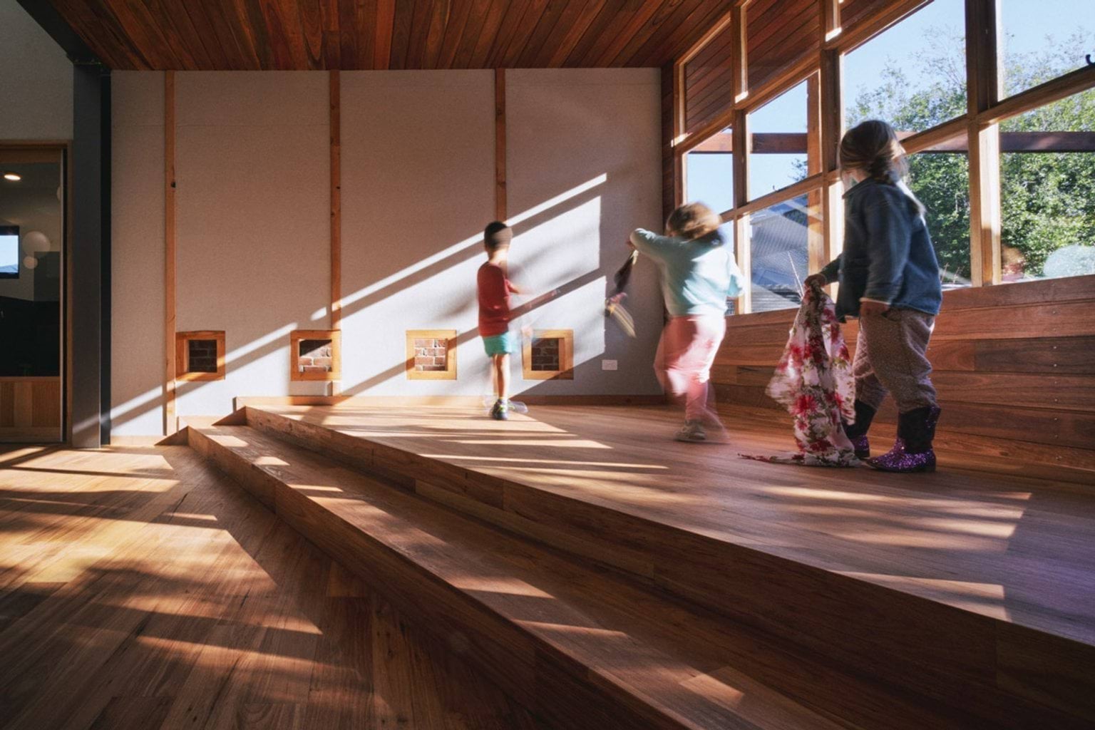Kids are standing and moving on a raised wooden floor inside St Kilda & Balaclava Kindergarten, with windows behind them. 