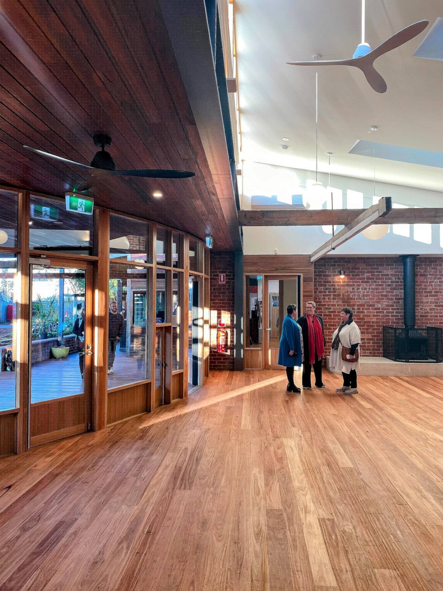 Inside St Kilda & Balaclava Kindergarten. Three adults are standing in a room with wooden floors. There is a brick wall behind them with a fireplace, and ceiling fans hanging from the high ceiling. There is a wall of windows and doors on the left showing the outside.
