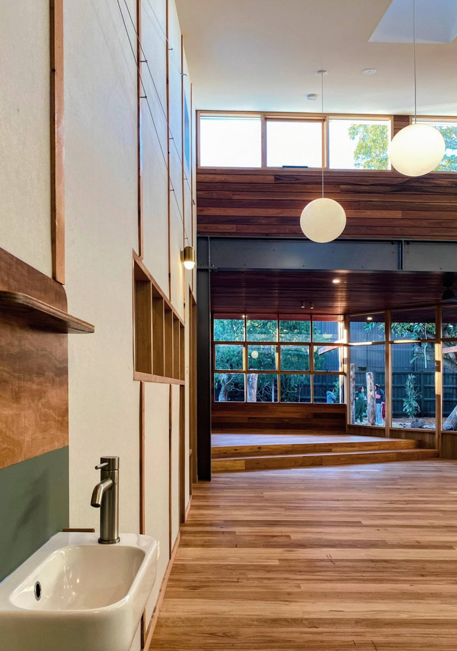 Inside St Kilda & Balaclava Kindergarten. There is a sink on the wall in the foreground and a wall of windows in the background. There are lights hanging from the ceiling. 
