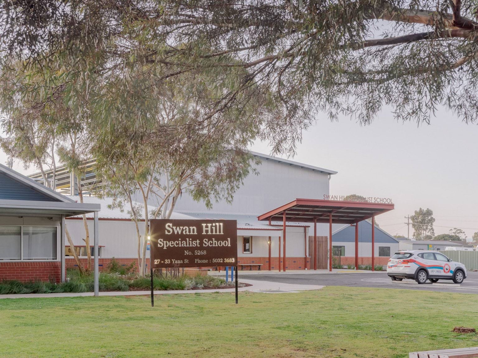 The entrance of Swan Hill Specialist School. There is a sign with the school name standing in the grass. Buildings are in the background with a road on the right for a drop-off area. 