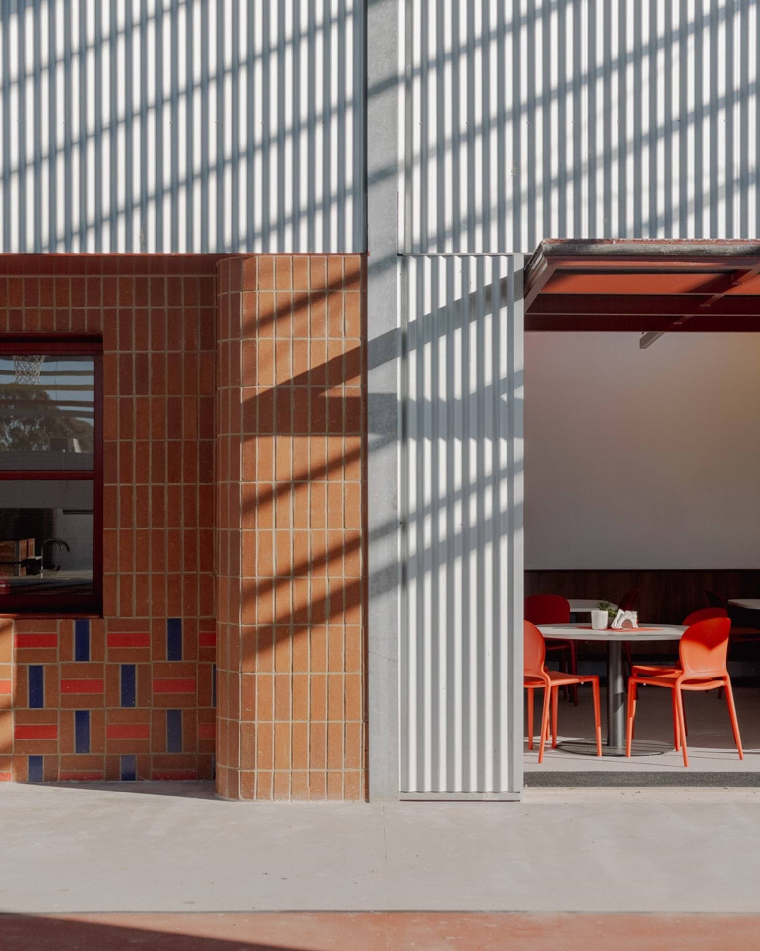 A close-up of the outside of a Swan Hill Specialist School building, showing a brick wall on the left next to a metal-clad wall on the right. The dining area with tables and red chairs is visible inside. 