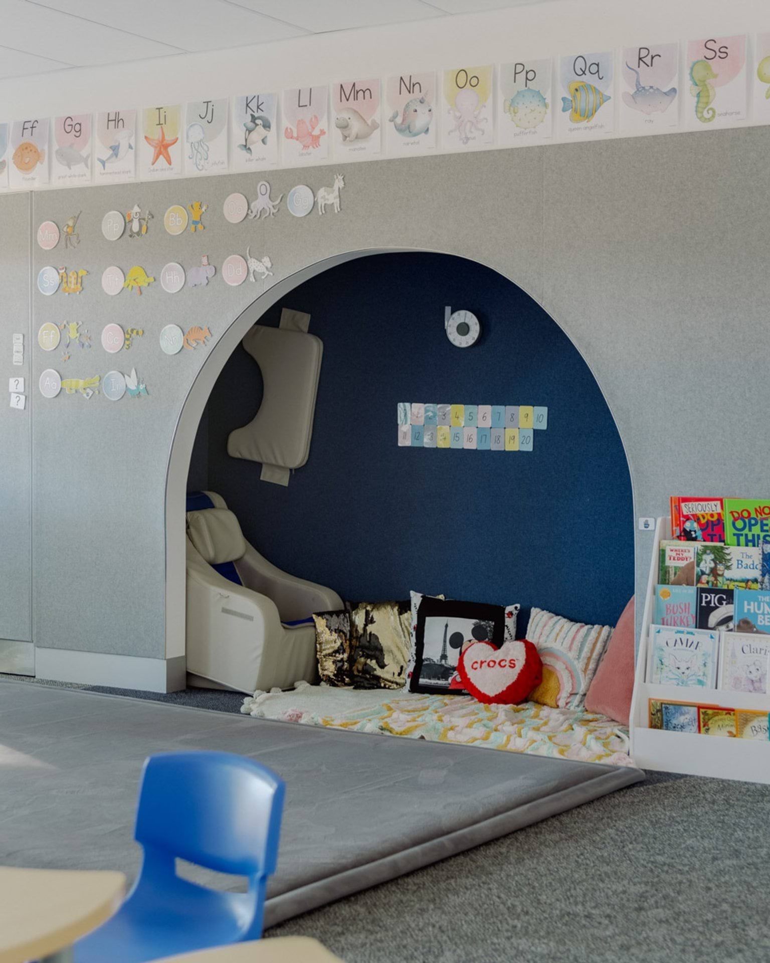 A classroom in Swan Hill Specialist School. There is a cut-out nook in a wall with pillows on the ground and an armchair. There are illustrations with the alphabet along the wall and picture books in a bookshelf.
