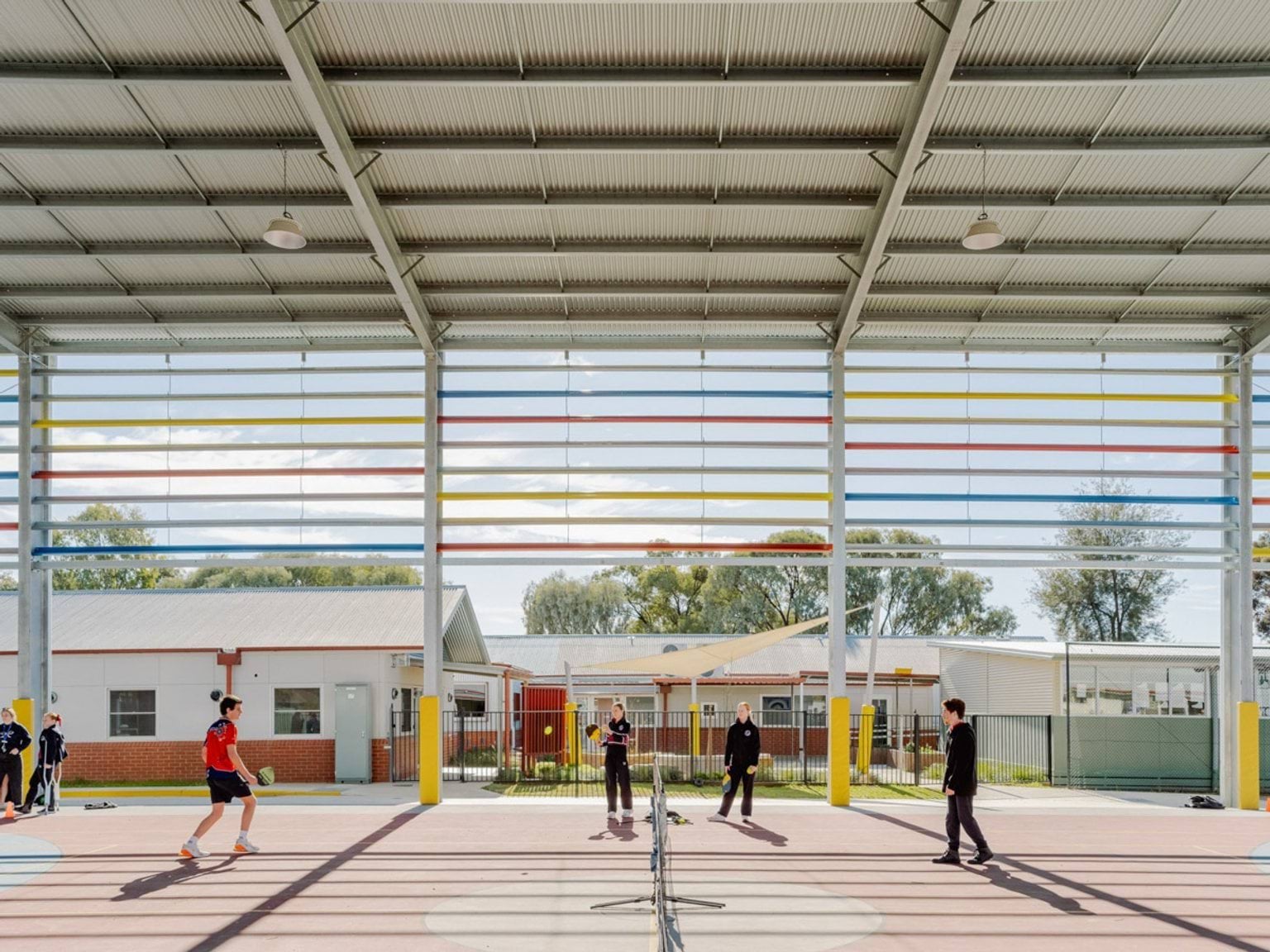 Another angle of the hard court inside the gymnaisum of Swan Hill Specialist School. The tennis net is in the middle with students on either side. The side of the gym behind them is open with coloured horizontal bars covering the top section.