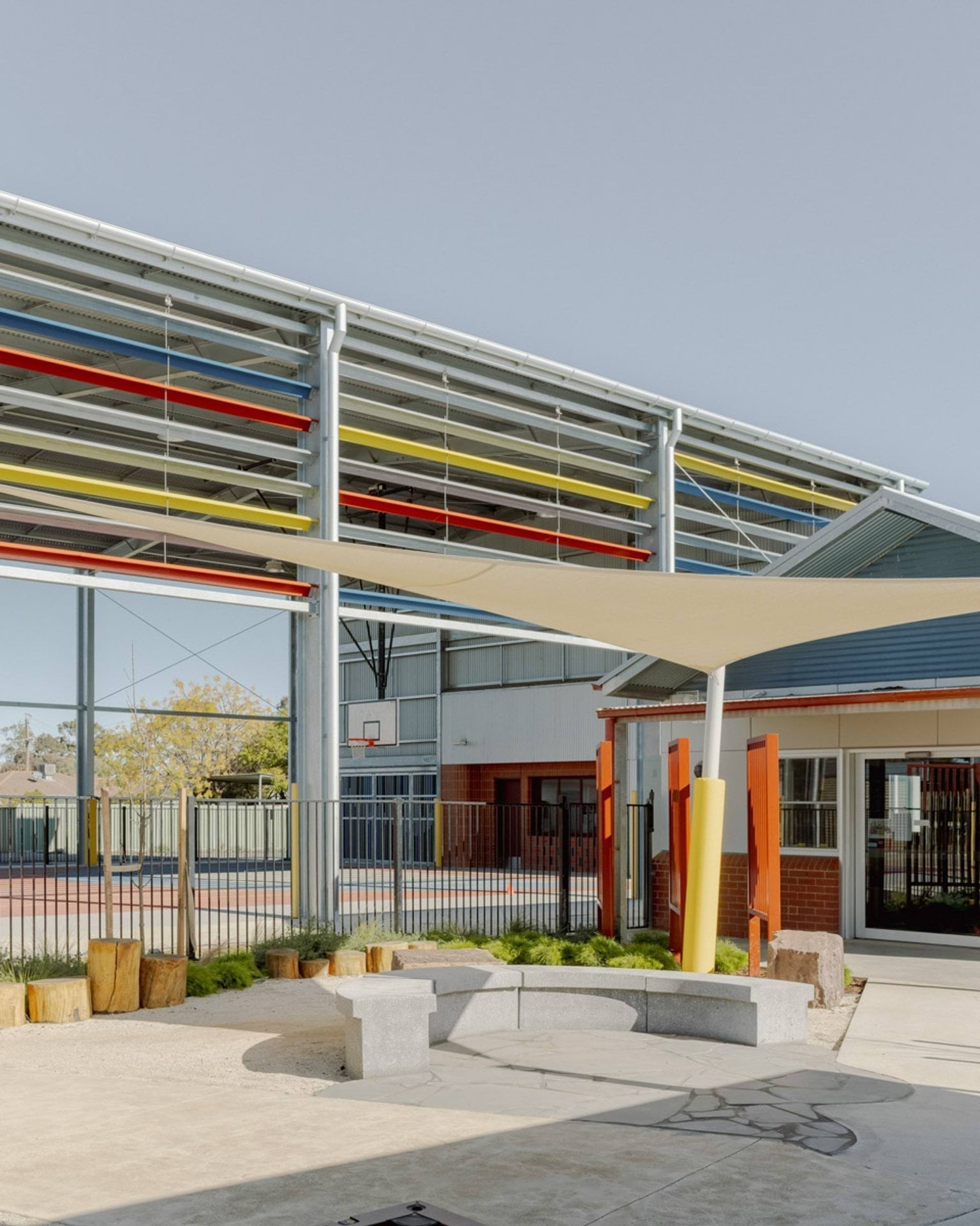 Outside Swan Hill Specialist School. There is a concrete curved bench to sit on with a shade sail above it. Behind is the gymnasium with the hard court.