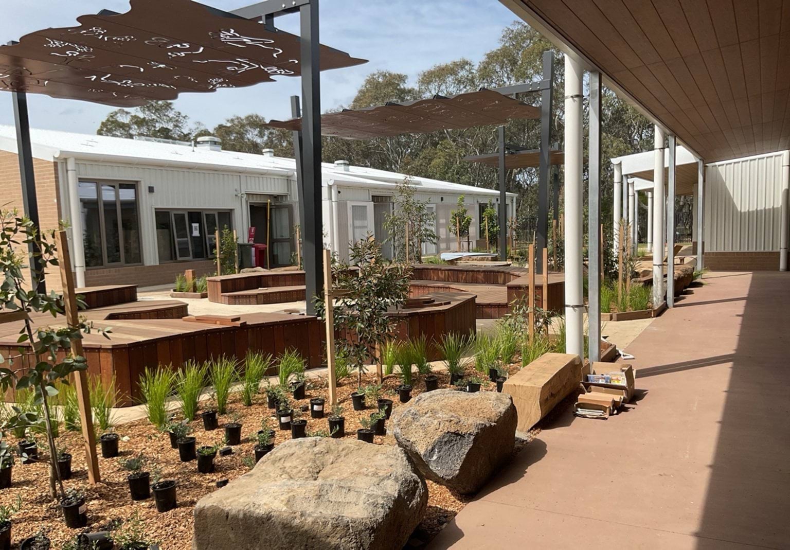 Biyala Primary School newly landscaped internal courtyard with timber and boulder seating. Newly planted native grasses and saplings and metallic sunshades continue to feature.