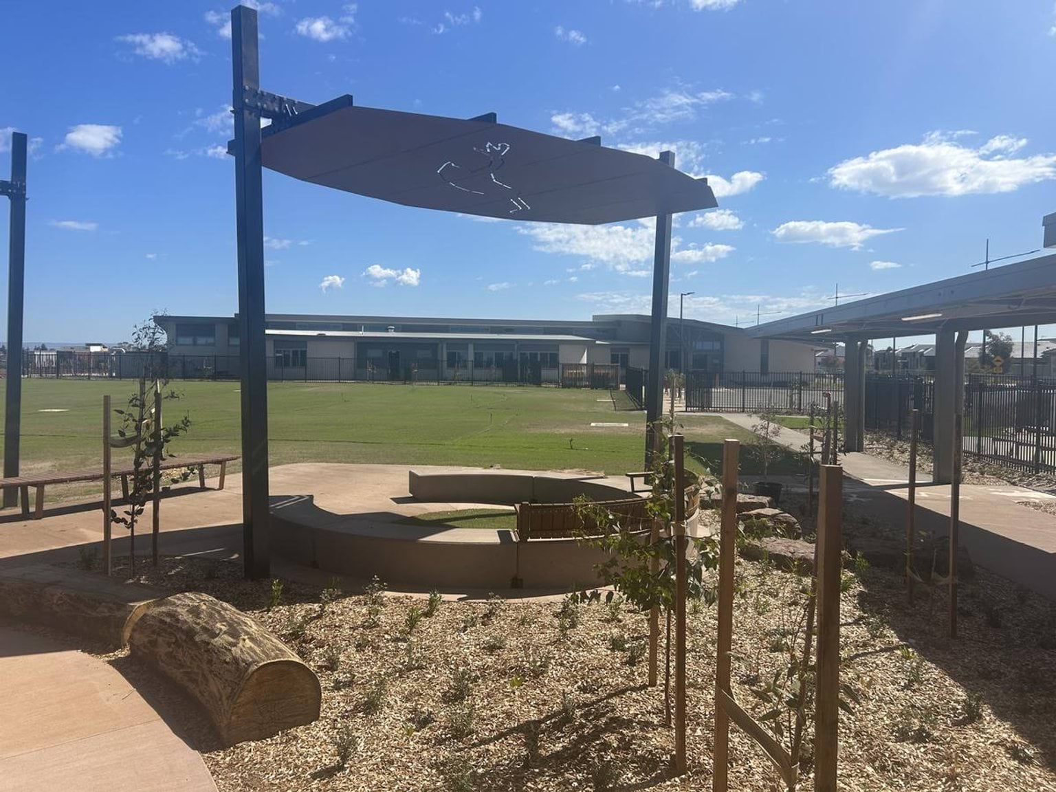 A photo of the outside area. There is a curved concrete seat with a shade sail above it. There are also new plants in the garden.