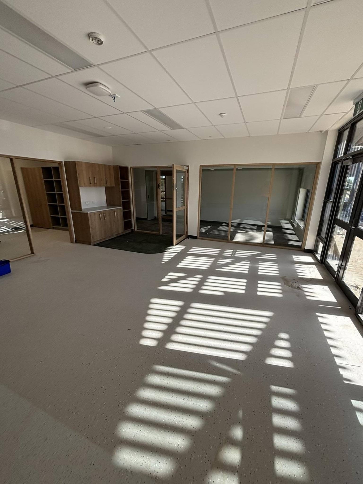 Inside the new Early Learning Victoria Numurkah (interim name) building, facing one side of the room that has a wall unit as well as glass doorways into other rooms. There are shadows on the ground from the sun coming through the windows and doors on the right. 