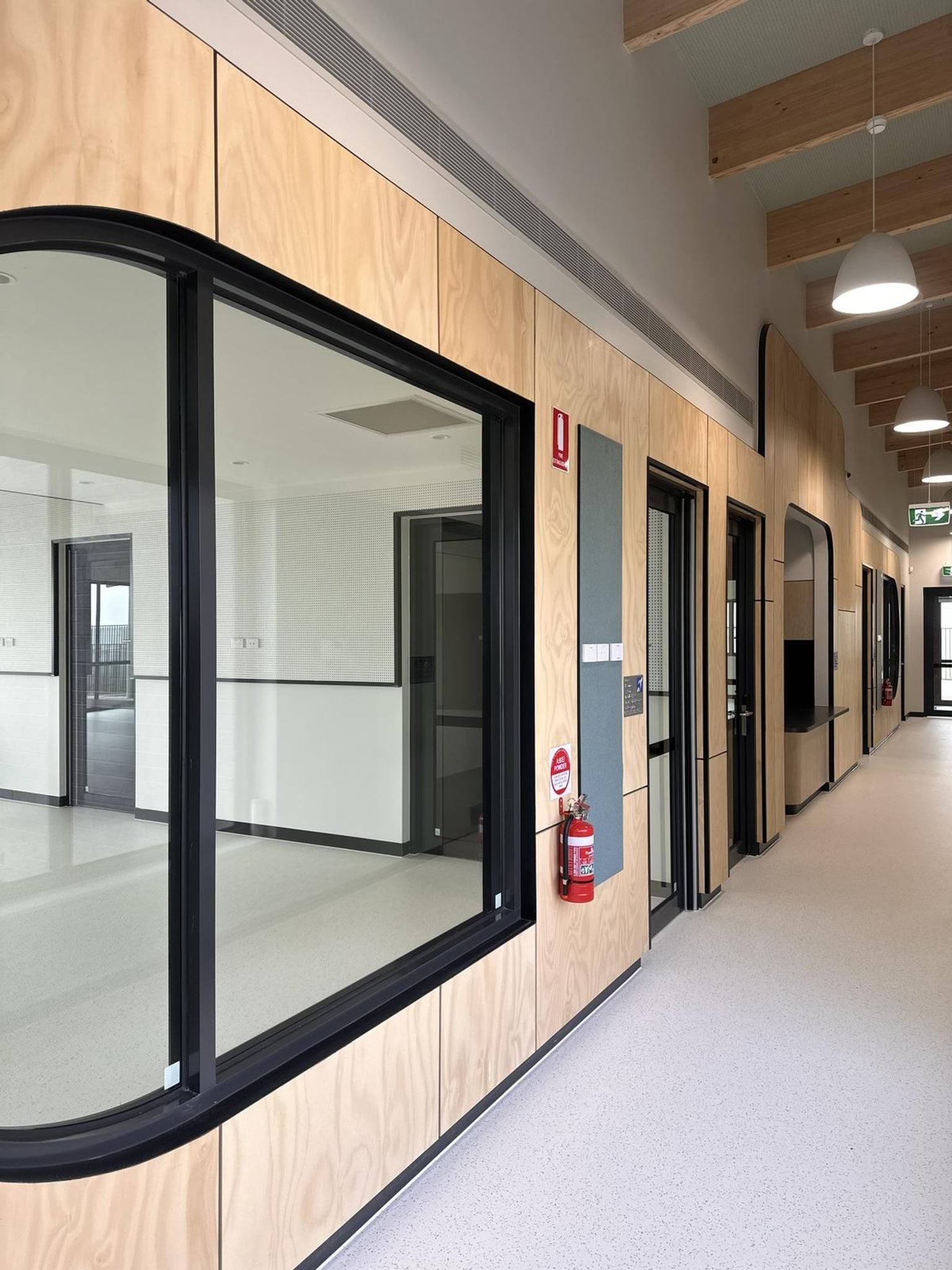 Inside the new Muyan Primary School Kindergarten (interim name) building, looking down a hallway with rooms on the left. Lamps hang from the ceiling that also has wooden rafters spaced evenly.