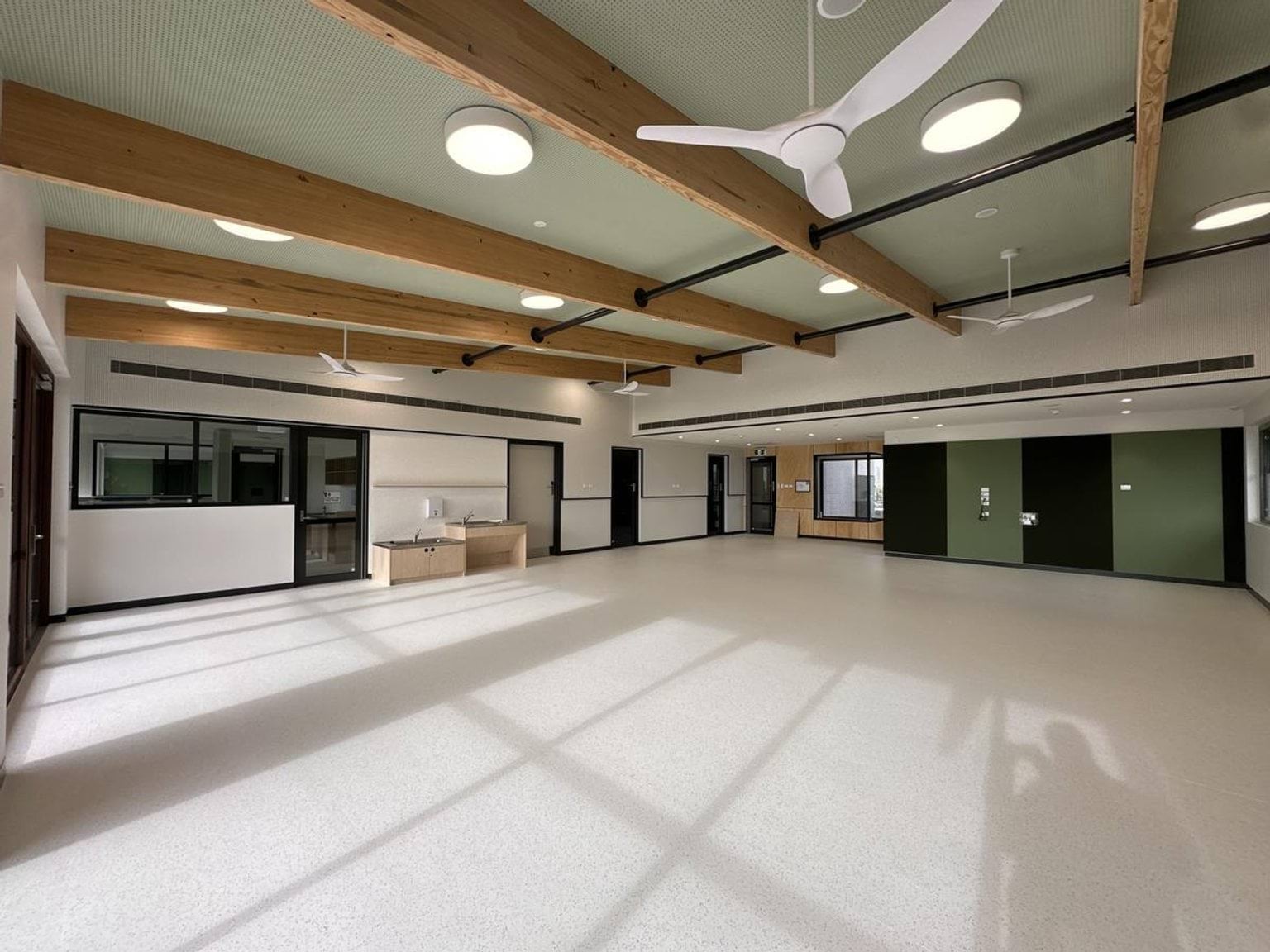 Inside the new Muyan Primary School Kindergarten (interim name) building, facing one corner. There are shadows on the ground from the sun coming through the windows behind the photo taker. There are fans hanging from the ceiling that also has wooden rafters spaced evenly. The room is mostly empty except for 2 sinks on a far wall.